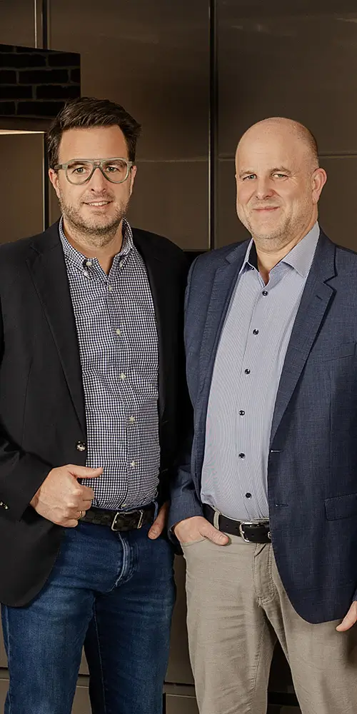 Wendlinger Mitarbeiter in Hemd und Jacket stehen vor einer modernen Küche mit dunklen Schränken und Backsteinwand.